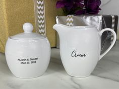 two white pitchers with names on them sitting next to a vase and flower pot in front of a gold box