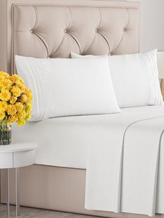 a bed with white sheets and yellow flowers in a vase on the nightstand next to it