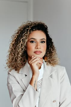 a woman with curly hair wearing a white jacket and gold earrings, posing for the camera