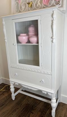 a white cabinet with glass doors and pink dishes on it's top, next to a wooden floor