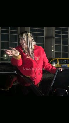 a woman in a red jacket standing next to a car with her arms outstretched out