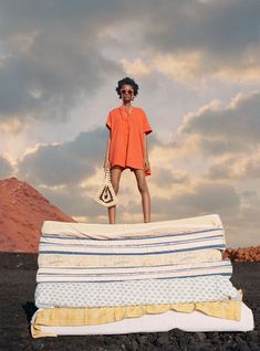 a woman in an orange shirt standing on top of a stack of mattresses with mountains in the background