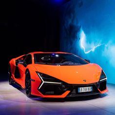 an orange sports car is on display in front of a blue wall with lightning behind it