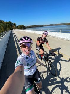 two bicyclists are riding down the road with their helmets on and one is taking a selfie