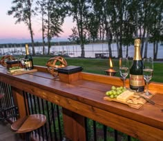 two bottles of wine and some cheese on a wooden table outside with water in the background