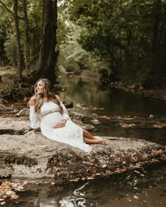 a pregnant woman sitting on top of a rock next to a stream in the woods