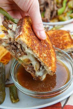 a hand holding a sandwich over a bowl of sauce and pickles on the side