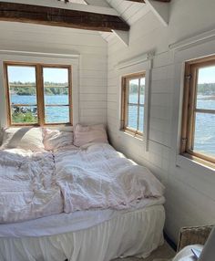 an unmade bed in the corner of a room with two windows and a view of water