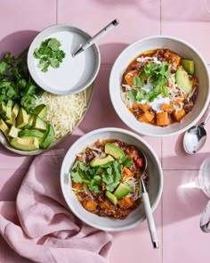 three bowls filled with food sitting on top of a pink tablecloth next to two spoons