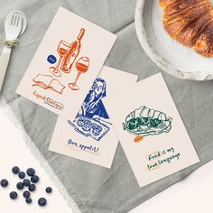 three cards with different illustrations on them next to a bowl of fruit and croissants