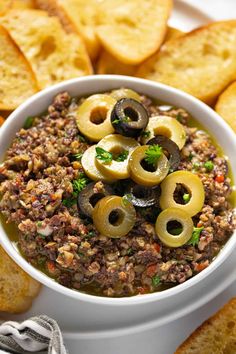 a white bowl filled with olives and ground beef dip surrounded by crackers on the side