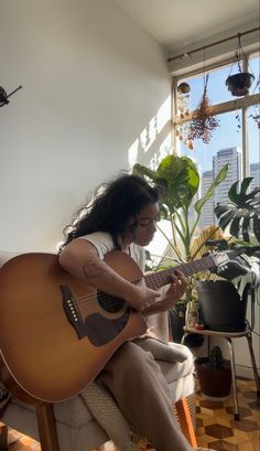 a woman sitting on a chair playing an acoustic guitar in front of a potted plant