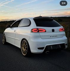 the rear end of a white car parked in front of a field at sunset or dawn