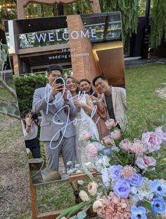 a group of people taking pictures in front of a welcome sign with flowers on it