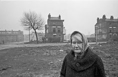 an old woman standing in the middle of a dirt field next to some buildings and trees