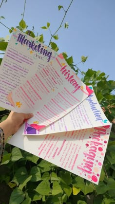 a person holding up three papers with writing on them in front of green plants and blue sky