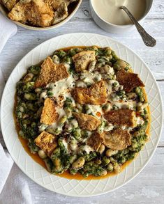 a white plate topped with food next to a bowl of soup and spoons on top of a wooden table