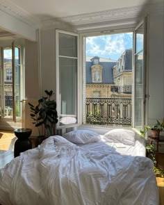 an unmade bed with white sheets and pillows in front of two large windows overlooking the city