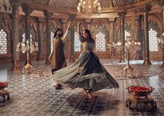 two women in long dresses are dancing inside an ornately decorated room with chandeliers