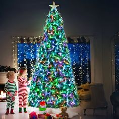 two small children standing in front of a christmas tree with lights and presents on it