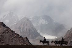 three people are riding horses in the mountains on a cloudy day with snow capped peaks behind them