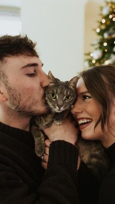 a man and woman kissing while holding a cat in front of a christmas tree with lights on it