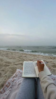 a person sitting on the beach with a book and pen in their lap looking at the ocean