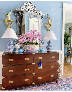 a dresser with flowers on top and two lamps next to it in front of a mirror