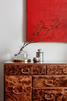 a painting hangs on the wall above a wooden dresser with drawers and brass handles, as well as a vase filled with flowers