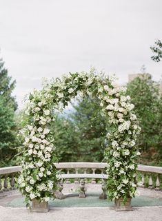 an arch covered in white flowers on top of a cement bench next to some trees