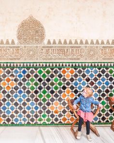 two small children sitting on wooden chairs in front of a wall with colorful tile designs