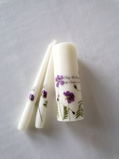 three white candles with purple flowers on them sitting next to each other, one is empty