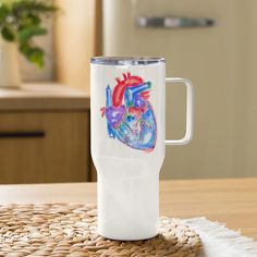 a white travel mug with a heart painted on the side sitting on a woven place mat