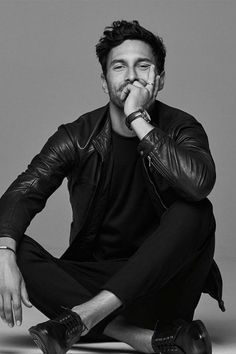 a man sitting on the ground with his hand to his mouth and wearing a leather jacket