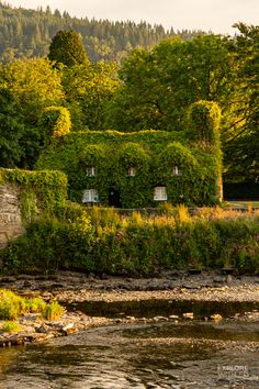 an old stone house sitting on the side of a river next to a lush green forest