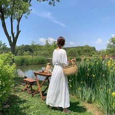 a woman in a white dress holding a basket next to a lake with yellow flowers