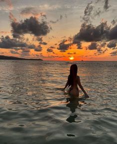 a woman is sitting in the water with her back to the camera as the sun sets