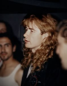 a man with long curly hair standing in front of other men and looking off into the distance