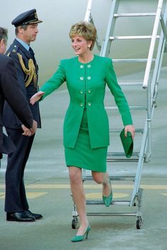 a woman in a green suit and hat is walking towards an airplane ladder as two men stand nearby