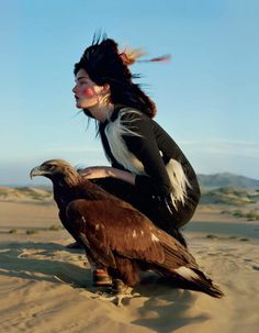 a woman sitting in the sand with an eagle on her lap