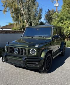 a green mercedes g - class is parked in front of a white wall and trees