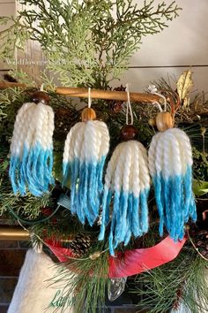 three white and blue knitted ornaments hanging from a christmas tree with pine branches in the background