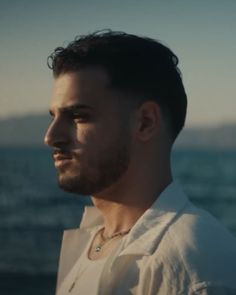 a man standing in front of the ocean wearing a white shirt and looking off into the distance