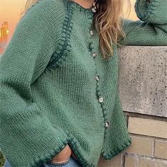 a woman wearing a green sweater and jeans leaning against a brick wall with her hands on her hips