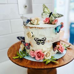 a cake decorated with flowers and butterflies on a wooden stand in front of a white tiled wall