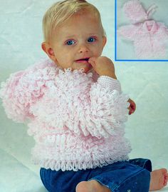 a baby sitting on the ground wearing a pink sweater and blue jeans with her hand under her chin