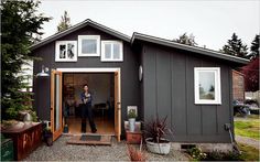 a man standing in the doorway of a small gray house next to trees and potted plants