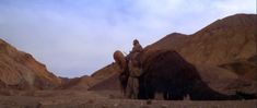 a man riding on the back of a brown horse in front of some rocks and mountains