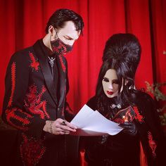 a man and woman dressed in black with red designs on their face, holding papers