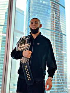 a man holding a wrestling belt in front of a large window with skyscrapers behind him